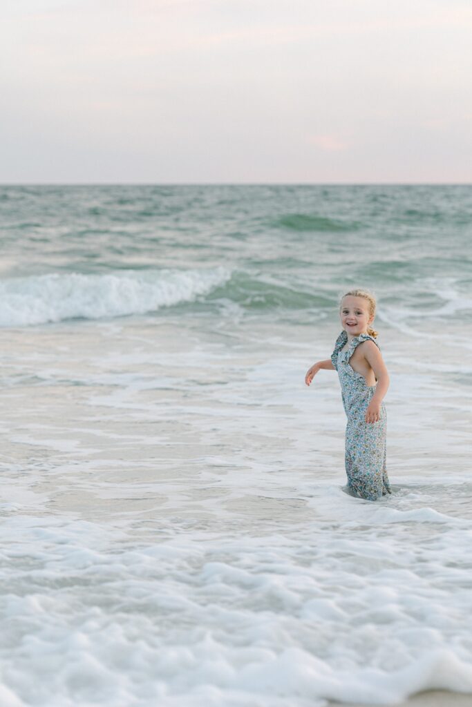 Young blond girl in a floral jumper, knee deep in the foamy ocean water, looking back toward camera and smiling.