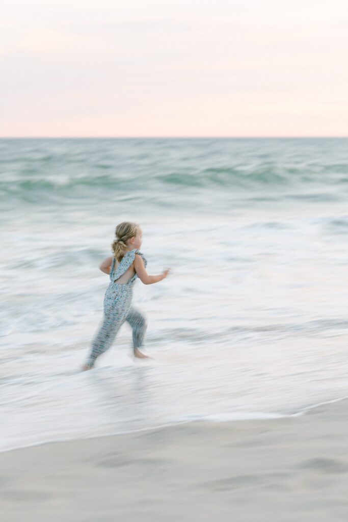 Blurry action shot of blond girl in floral jumper running with the waves of the turqoise ocean against the pink sky.