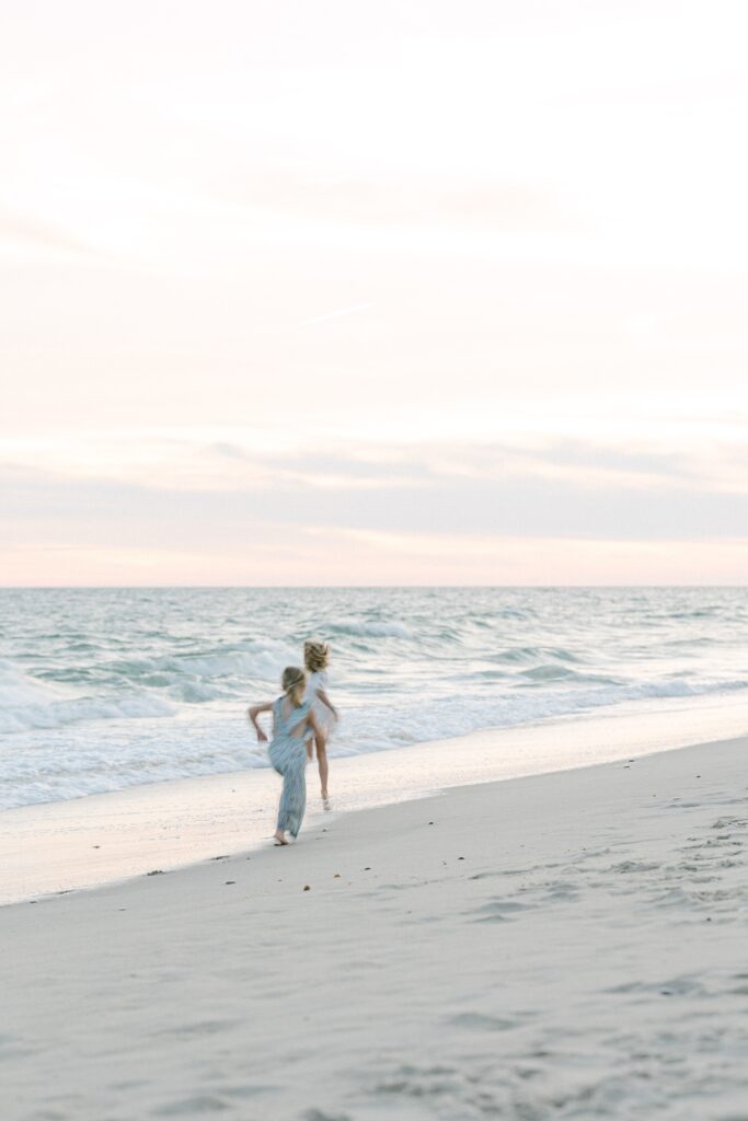 Two blonde sisters running along the ocean line in the sand. 
