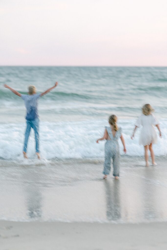 Blurry image of blond children playing near the ocean's edge - the oldest boy has both arms in the air in excitement and the blond sisters are walking toward the water. 