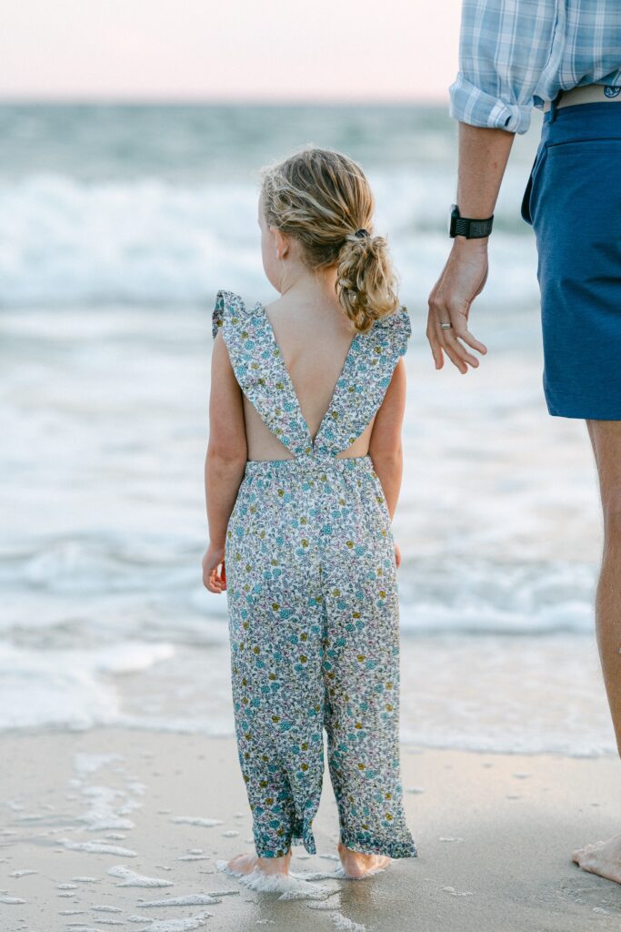 Young blond girl with curly hair in ponytail, wearing floral jumper and standing next to her father at the ocean's edge; shot from behind.