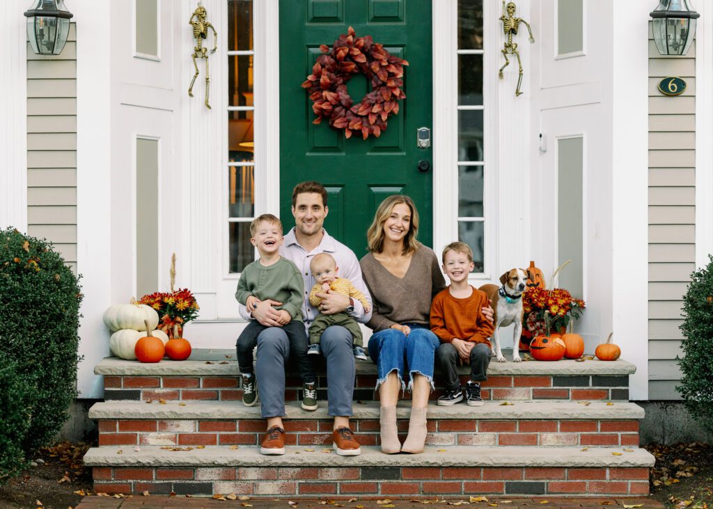 Family of 5 sitting on the front steps of their home, all in fall colors surrounded by pumpkins & mums with the dog standing before them. 