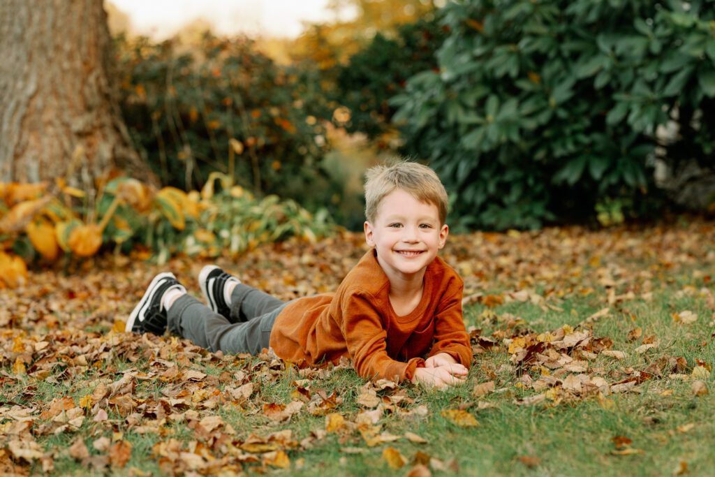 Older brother in rust colored shirt is laying on his belly in the leaves and grass, propping himself up on this elbows and smiling at the camera. 