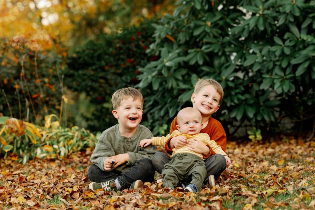Two brothers sitting in a bed of leaves criss cross applesauce smiling at the camera - the older has baby brother in his lap. 