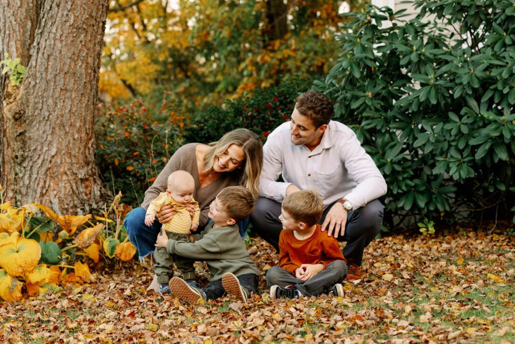 Family of 5 sitting in a bed of leaves on the grass, all eyes on baby boy who mom is holding up in a standing position. 