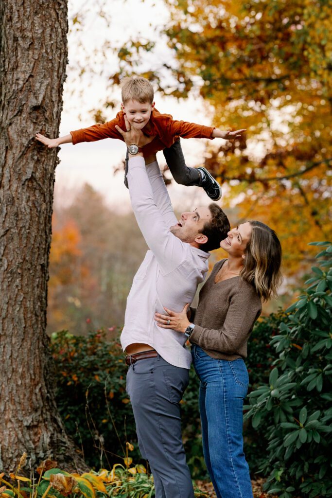 Dad holding oldest son up in the air who is in a flying superman pose, with mom standing behind dad resting her hands on his waist, smiling up at her boy. 
