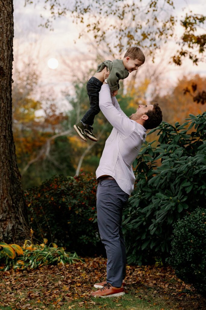 Action shot of dad about to throw toddler boy in olive shirt up in the air - son is smiling big down at dad. 
