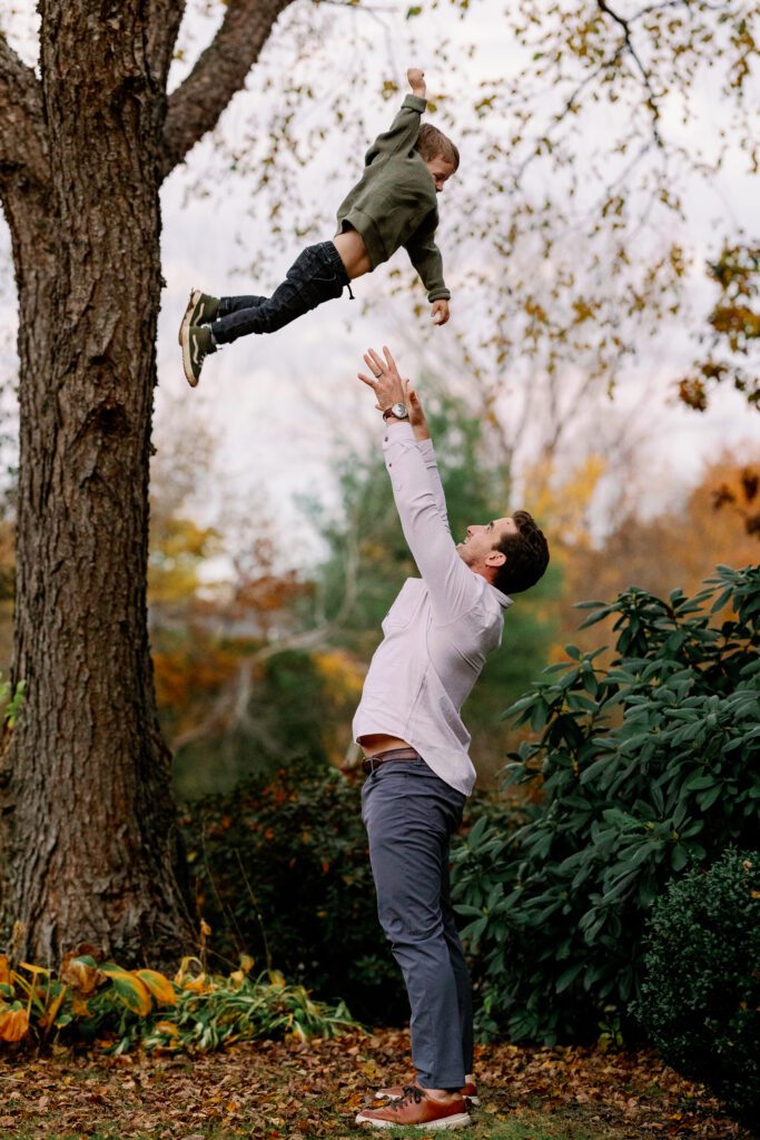 Action shot of dad having just thrown toddler boy in olive shirt up in the air - boy has arm raised in a superman pose and dad has arms outstretched preparing to catch him. 
