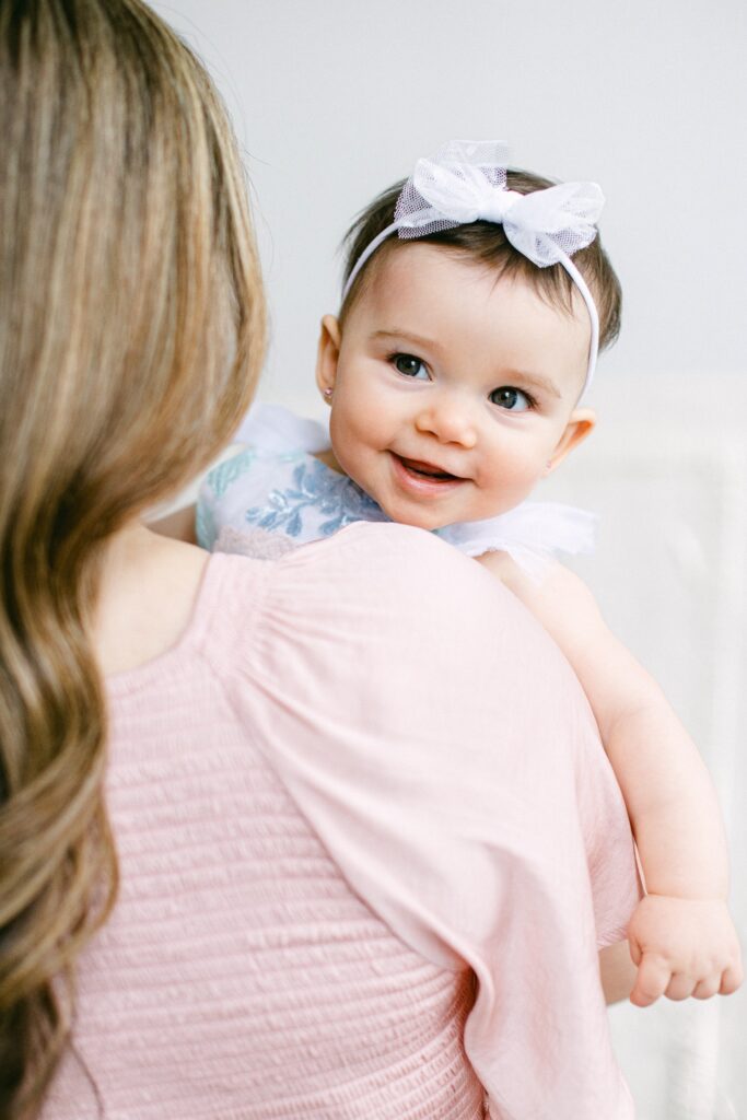 Baby girl wearing a white tulle bow being held by mom and smiling over her shoulder.
