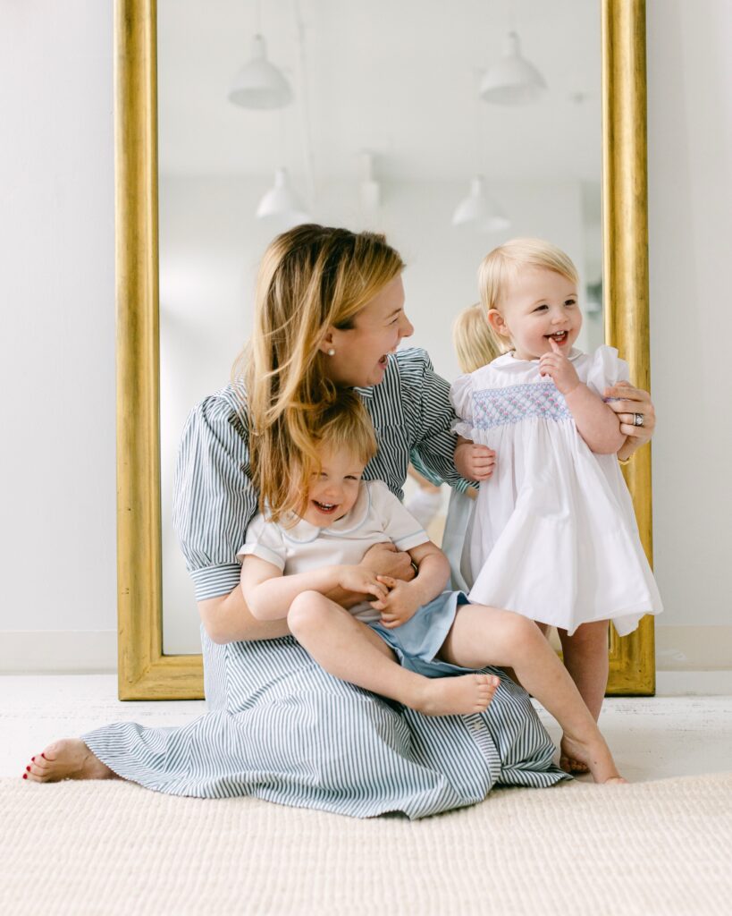 Mom is sitting in front of the mirror with one arm wrapped around the son sitting on her lap and the other around her daughter standing beside her and smiling off to the side with finger in mouth. 