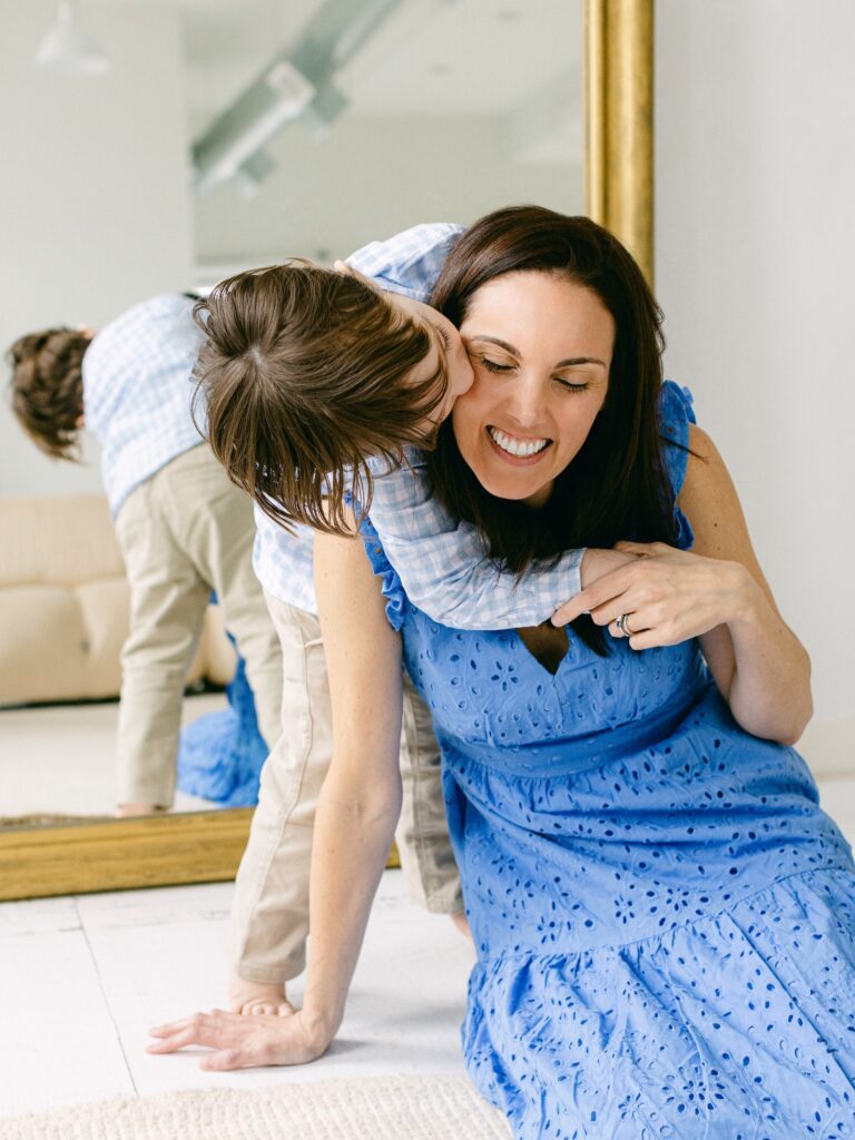 Mom in blue dress, sitting in front of a gold-framed mirror, gets a kiss on the cheek from her son in a blue and white checkered shirt and khaki pants.