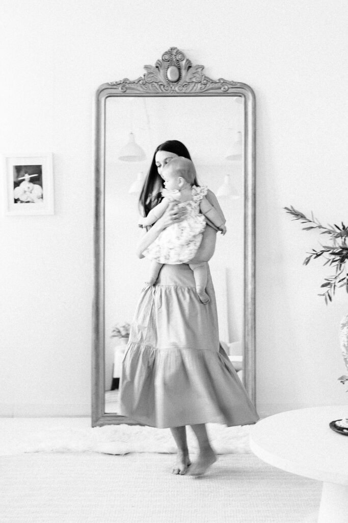 Black and white image of mom twirling in front of a large full-length mirror with baby in her arms.