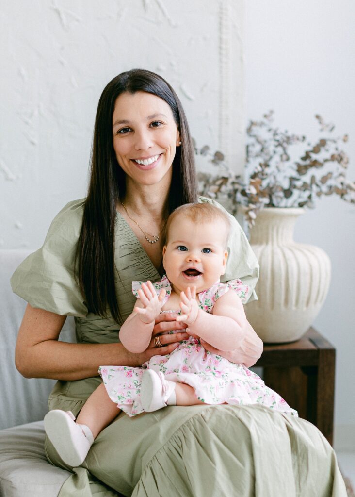 Smiling mom is sitting on a chair holding baby daughter who is mid-clap and smiling. 