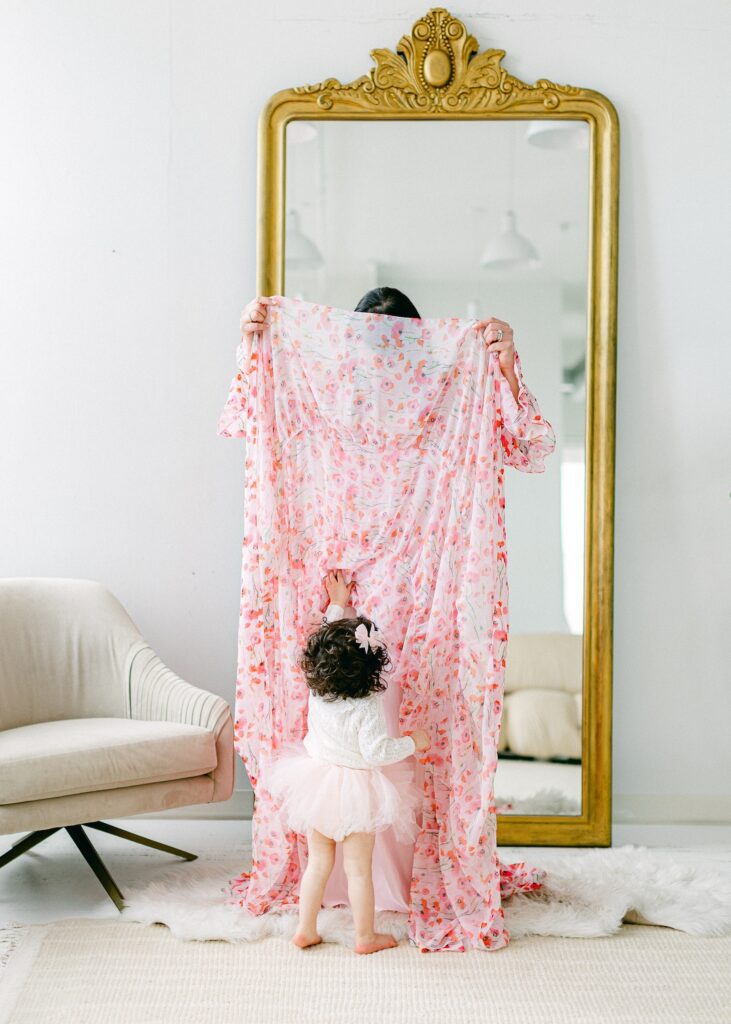 Mom is holding a layer of her flowy dress up in the air and hiding behind it, while daughter is standing in front of her and reaching up to pull the dress down. 