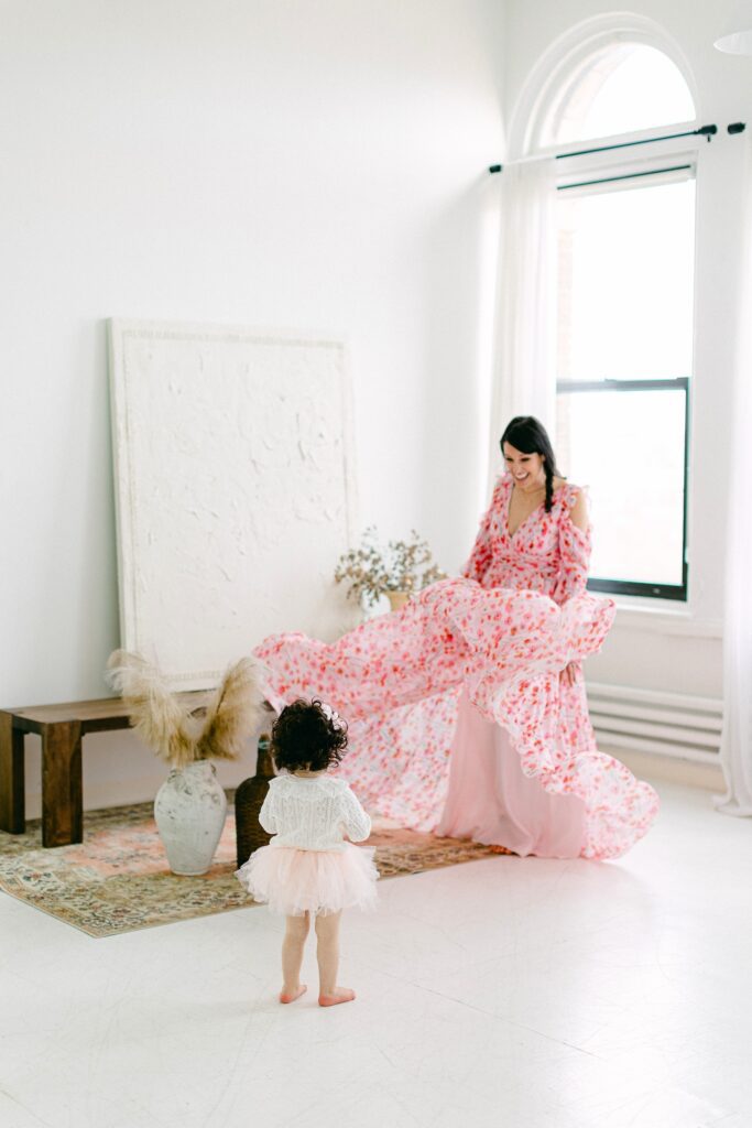 Mom is smiling at daughter and twirling flows dress while daughter watches on in amazement. 