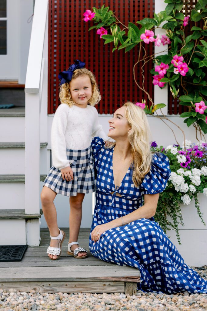 Mom in a blue and white plaid dress sitting on a wooden step smiling up at her daughter who is standing beside her smiling straight ahead.