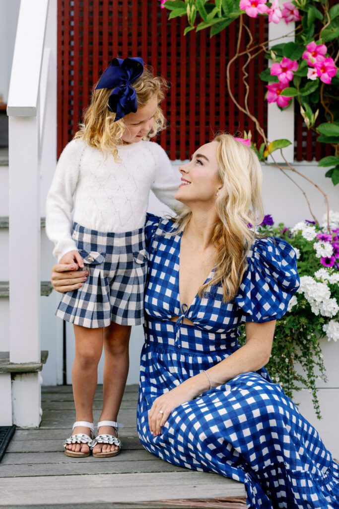 Mom in a blue and white plaid dress sitting on a wooden step smiling up at her daughter who is standing beside her looking into her eyes. 