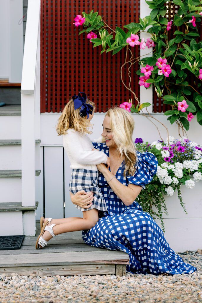 Mom in a blue and white plaid dress sitting on a wooden step holding daughter in her arm with both looking at something on the daughter's sweater.