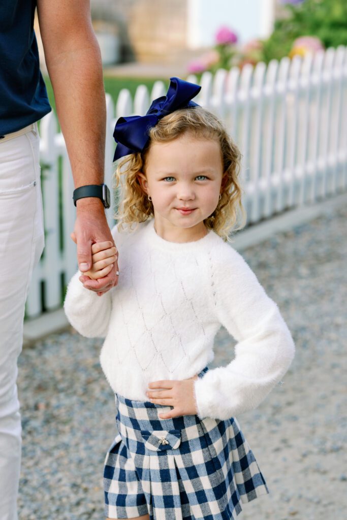 Daughter with big navy bow in her hair is holding dad's hand while her other hand is on her hip in a sassy pose with a little smirk on her face. 