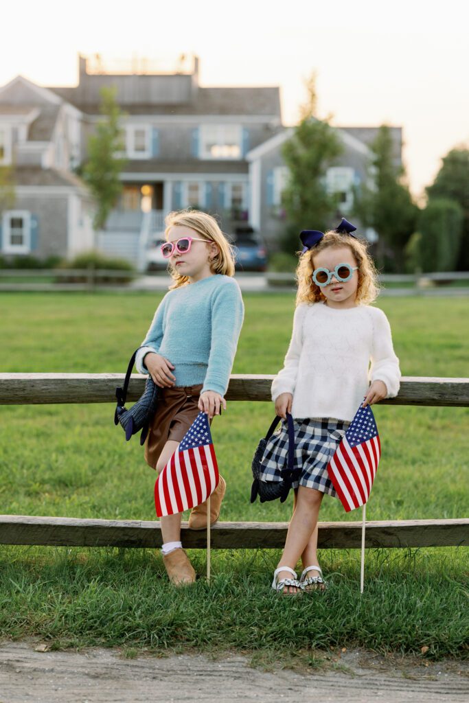 Both daughters are standing alongside a fence with colorful sunglasses on and holding navy purchased shaped like whales and American flags. 