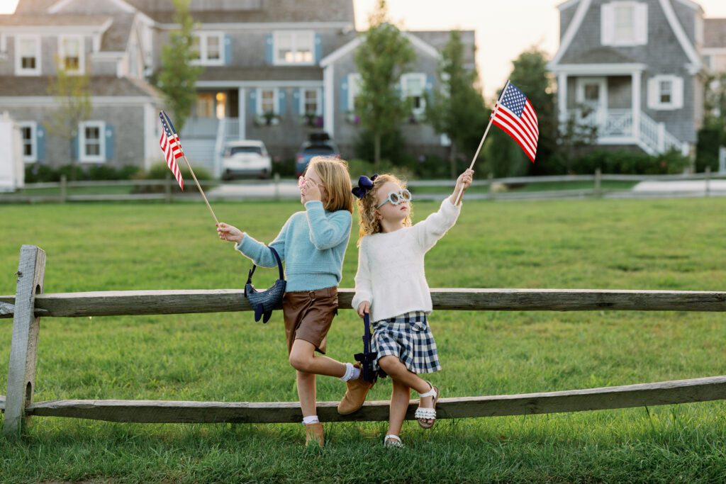 Both daughters are standing alongside a fence with colorful sunglasses on and holding navy purchased shaped like whales and American flags up in the air. 