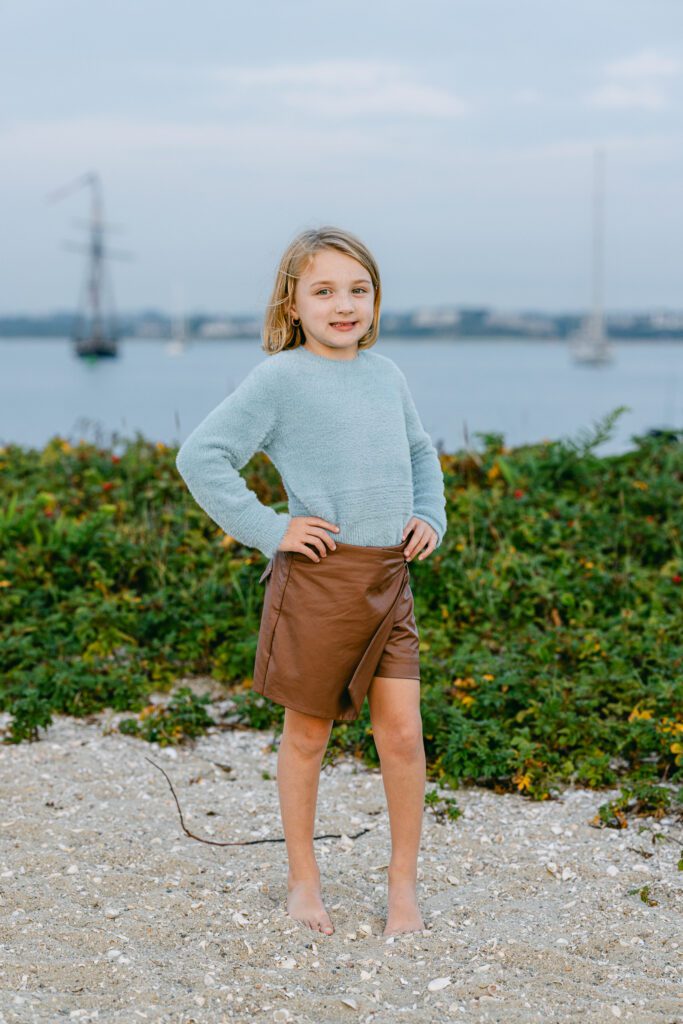 Daughter is standing with hands on her hips and smiling, the water and boats in the background. 