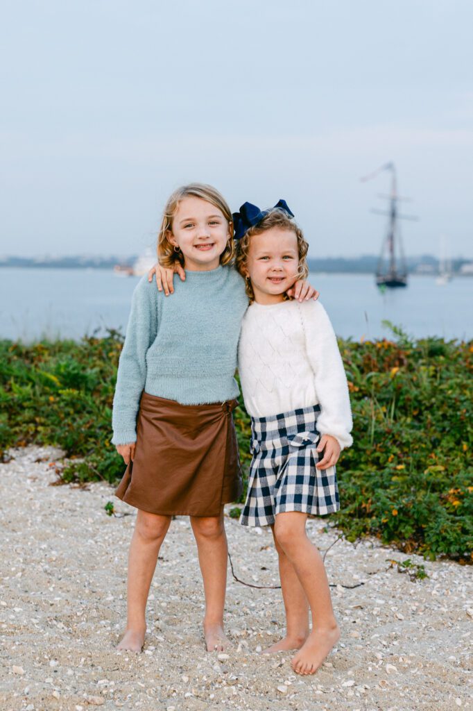 Sisters are standing side by side with the water and boats in the background, their arms around one another and smiling.