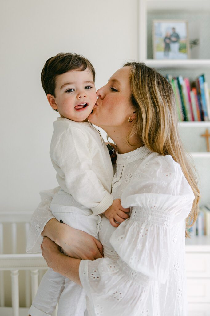 Mom is holding her toddler son and kissing his cheek, while the toddler son is smiling straight ahead with his tongue slightly hanging off to the side. 