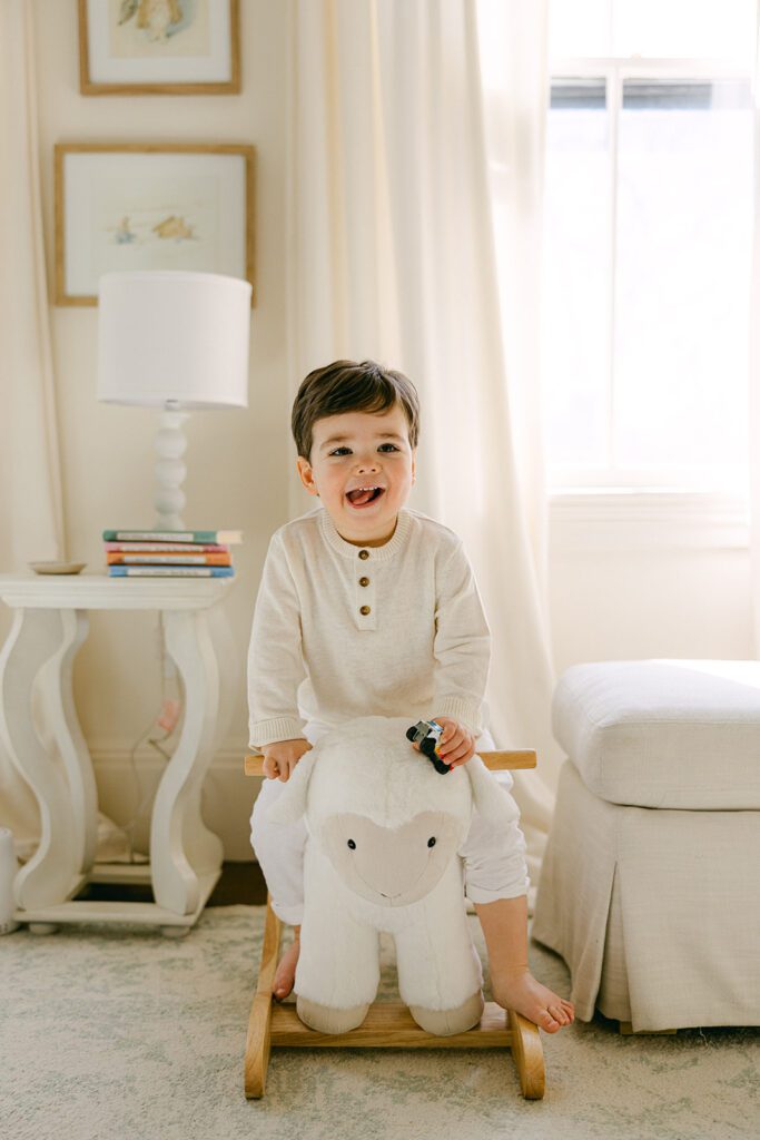 Toddler boy sitting on a white lamb rocking horse and smiling with his tongue slightly sticking out to the left. 