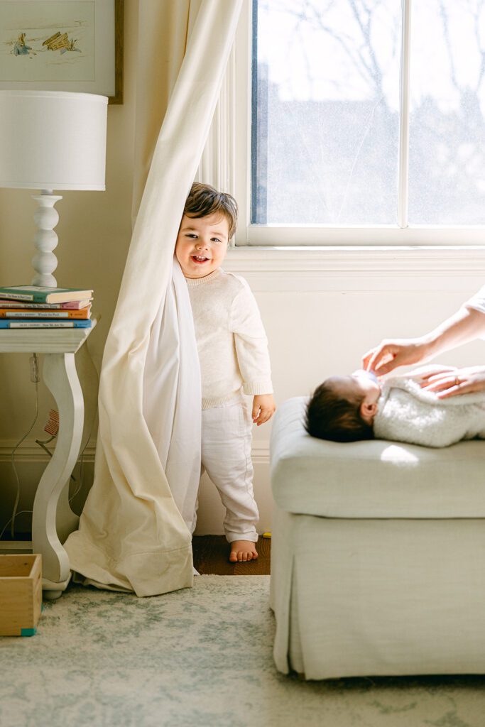 Toddler boy is standing behind the curtain is smiling while his newborn brother is getting help with a pacifier while swaddled on the cushioned ottoman. 