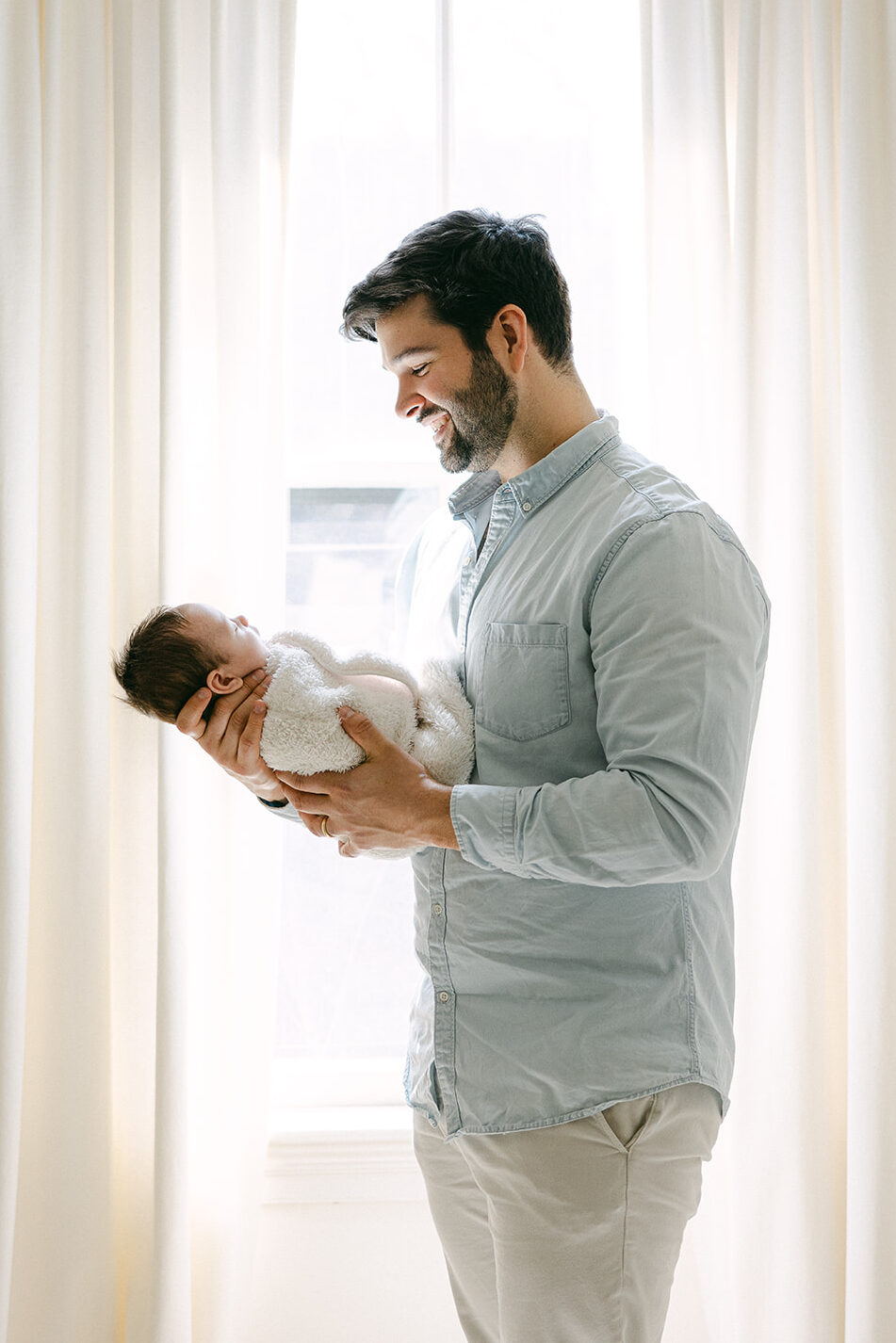 Dad is standing in front of a window with flow-y drapes and holding his newborn son out in front of him and smiling at him. 