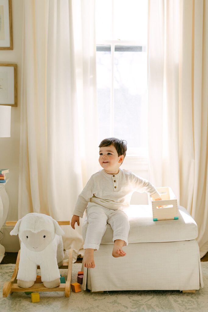 Toddler boy is sitting on a cushioned ottoman with one hand reaching for a block in his box while smiling off in the opposite direction. 
