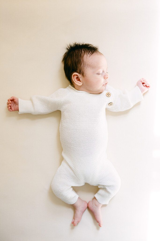 Newborn baby in ivory sweater outfit with arms spread out and sleeping. 