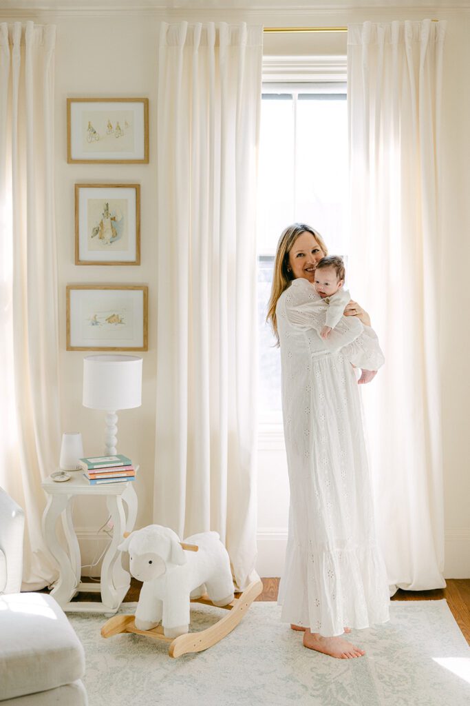 Mom holding newborn son in front of a bright window with flowing drapes and smiling. 