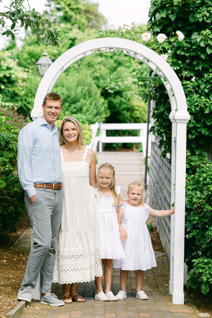 Dad, Mom, and two blonde sisters are standing in a row on a brick path under a white arch, arms around each other and smiling. 