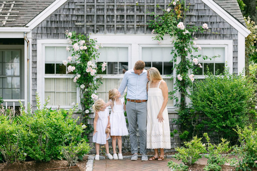 Two girls, Dad, and Mom are standing in a row in front of a gray shingled house with two big windows and climbing roses, all looking at each other. 