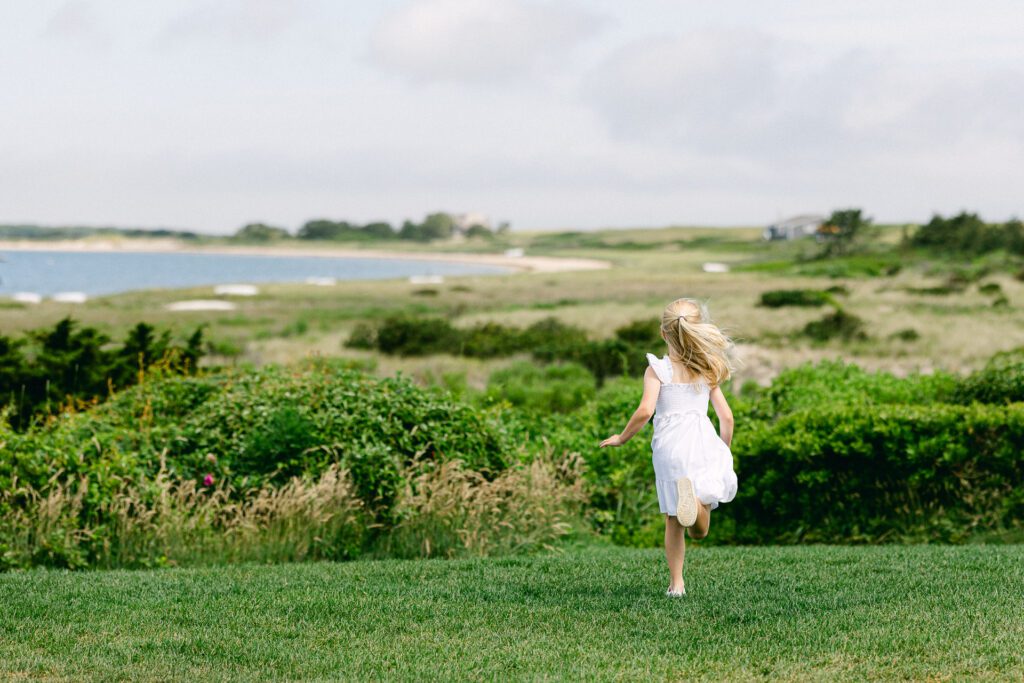 Blond girl in white dress is running on an elevated grassy patch, with clouds and ocean visible in the distance.