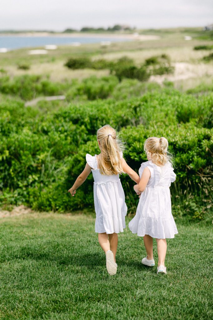 Blond sisters in white dresses holding hands and walking across a grassy area with the ocean visible in the distance. 