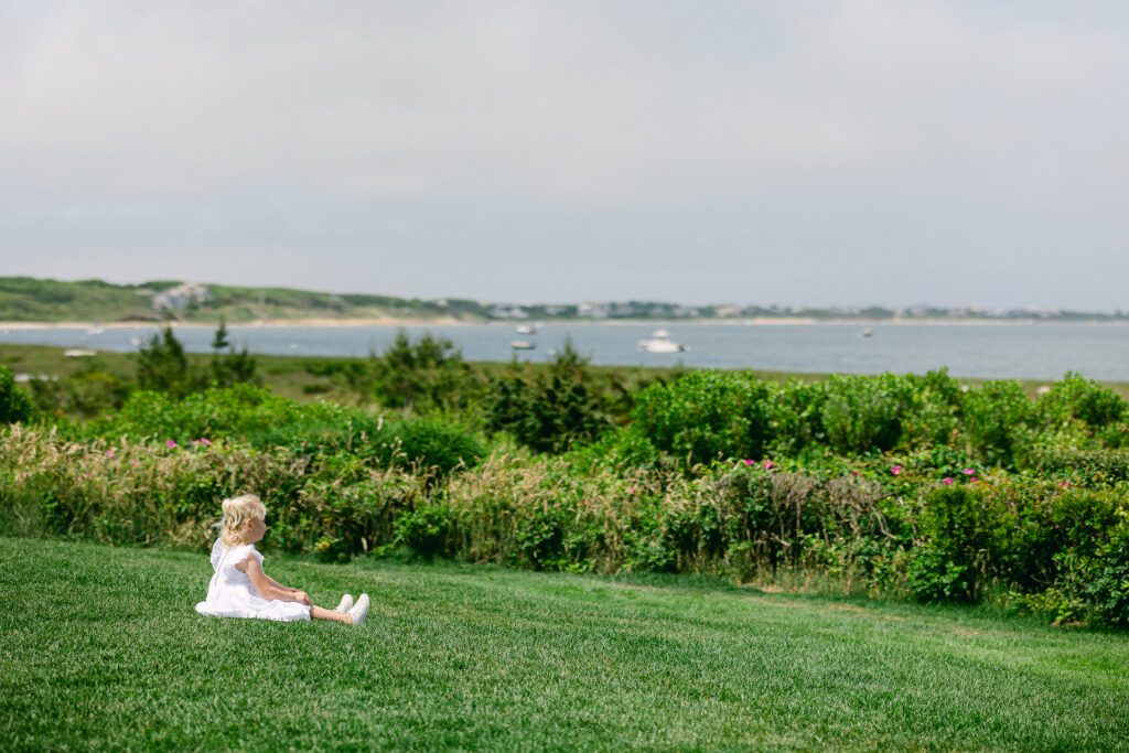 Young blond girl in white dress sitting on a lush green grassy lawn looking out at the ocean 