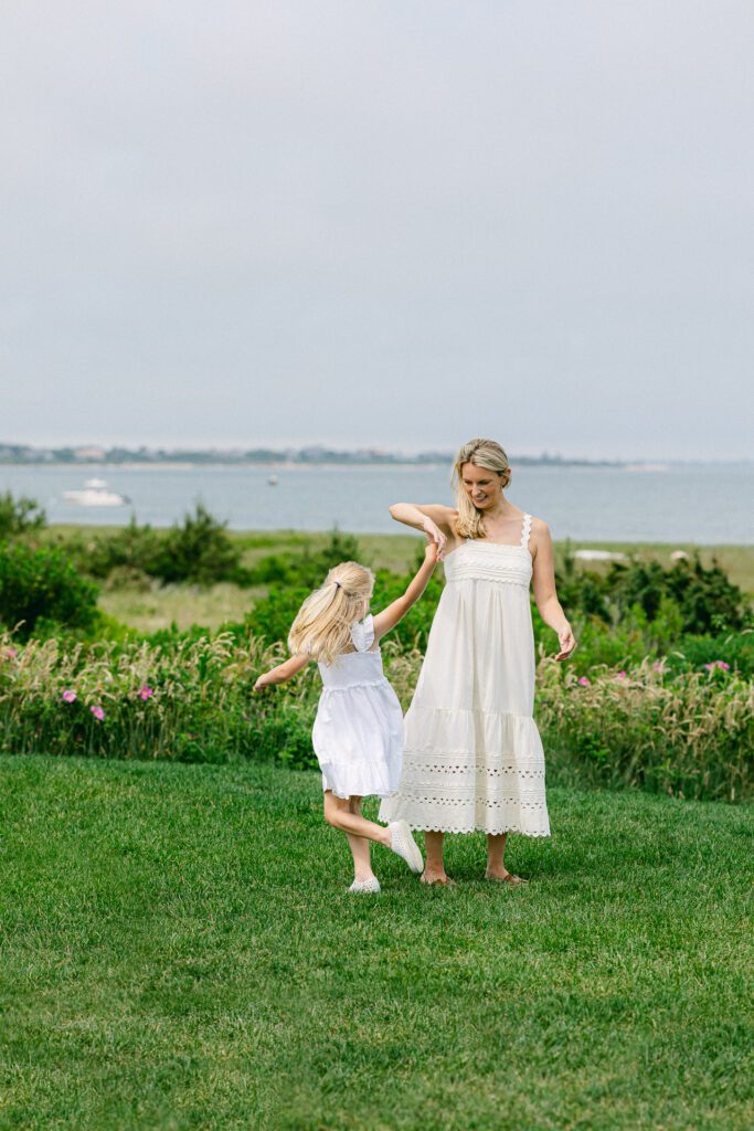 Mom is holding daughter's hand and twirling her in a circle on the green lawn in front of the ocean. 