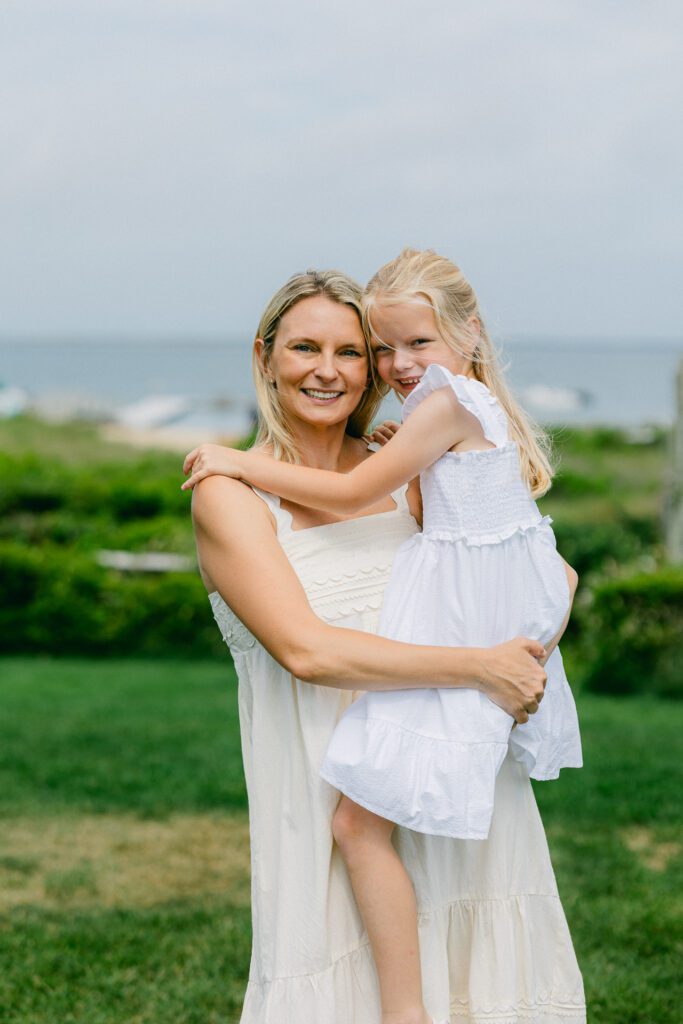 Mom and daughter in white dresses, standing on grass, with ocean in background. Mom is holding daughter on her hip and both are smiling. 