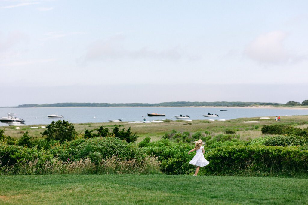 Blonde daughter in white dress is dancing on an elevated grassy area. Ocean and clouds are visible in the distance.