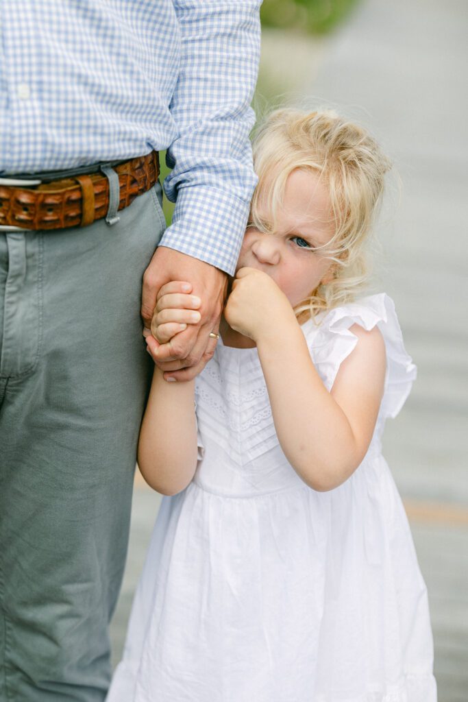 Young blond girl in white dress stands next to her father, one hand in her mouth and the other clutching dad's hand. 