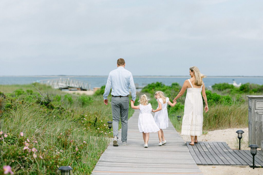 Family of four holding hands and walking down the boardwalk towards the beach. Mom and daughters wear white dresses.