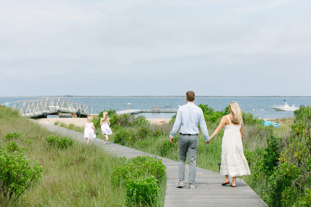 Dad and Mom holding hands and walking down the boardwalk towards the beach with their two blond daughters in white dresses running ahead of them. 