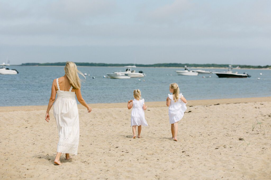 Mom and daughters with blond hair and white dresses all walking on the beach towards the water's edge with boats anchored in the water beyond. 