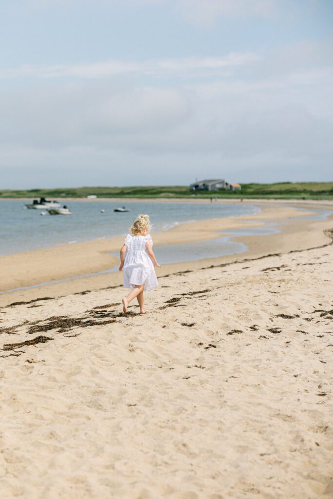 Blond toddler girl in white dress walking along the seaweed line on the beach. 