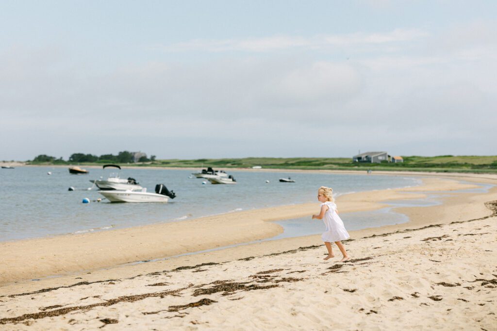 Blonde toddler girl in a white dress running on the beach towards the water. 