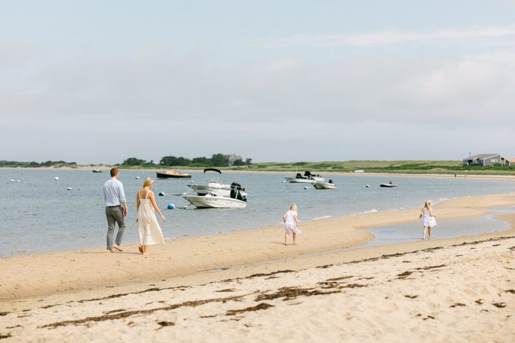 Dad, Mom, and two sisters in white dresses walking around down the beach with boats anchored beyond in the water. 