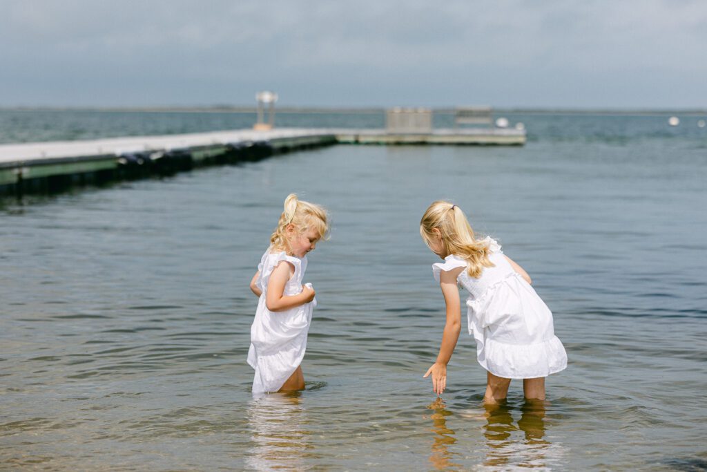 Two blonde girls in white dresses wading in the shallow water of the ocean tide, with the oldest girl dipping her hand in the water. 